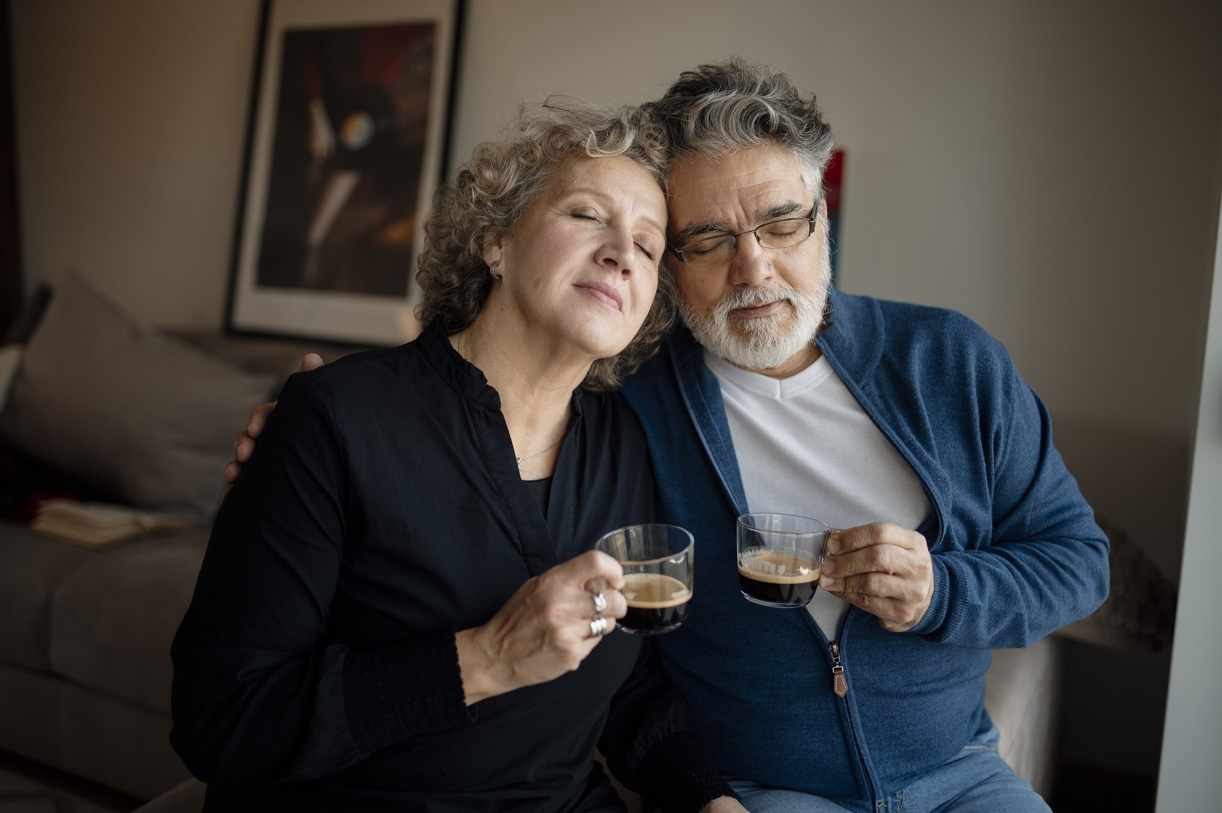 Een ouder echtpaar zit dicht tegen elkaar aan in de woonkamer, elk met een kopje koffie.