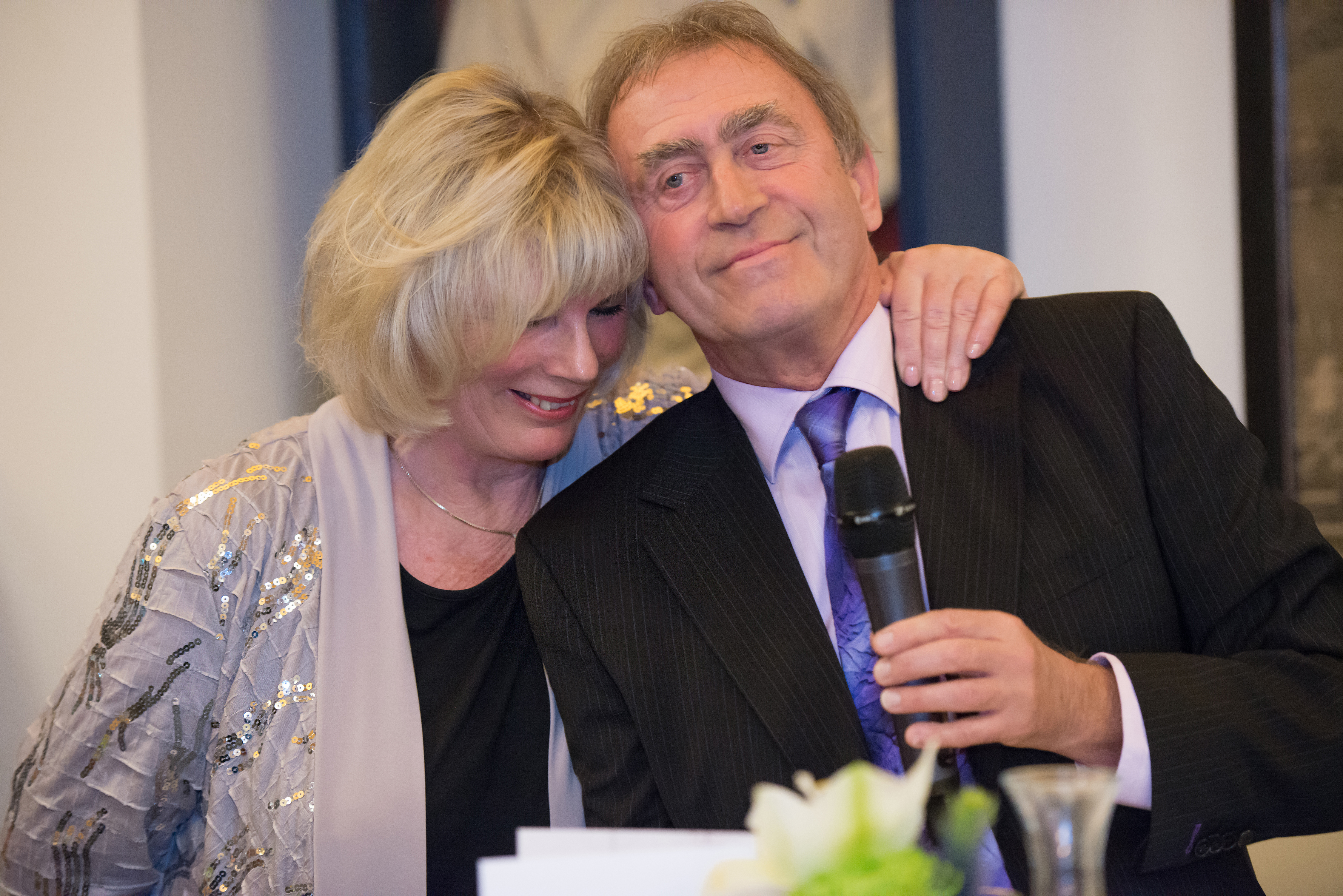Ed van Peppen lacht met een microfoon in de hand, terwijl een vrouw een arm om hem heen slaat.