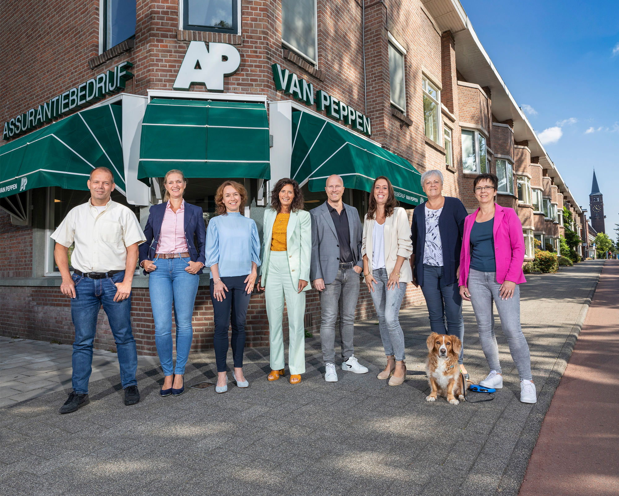 Het team van Van Peppen, acht mensen en de kantoorhond, op de stoep voor het hoekpand met de groene markiezen en de letters AP en Van Peppen.