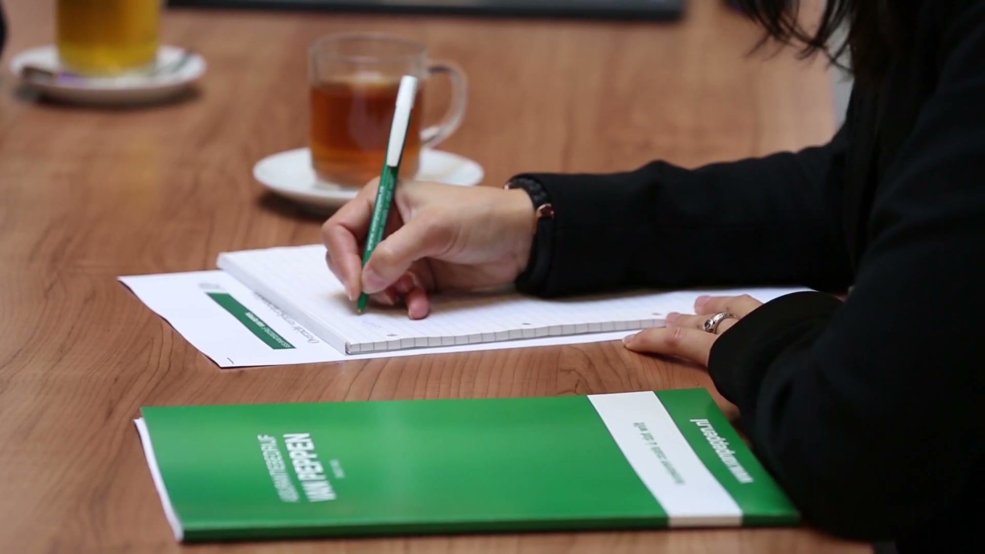 Close-up van een hand die met een groene pen schrijft, naast een groene map van Van Peppen en een glas thee.