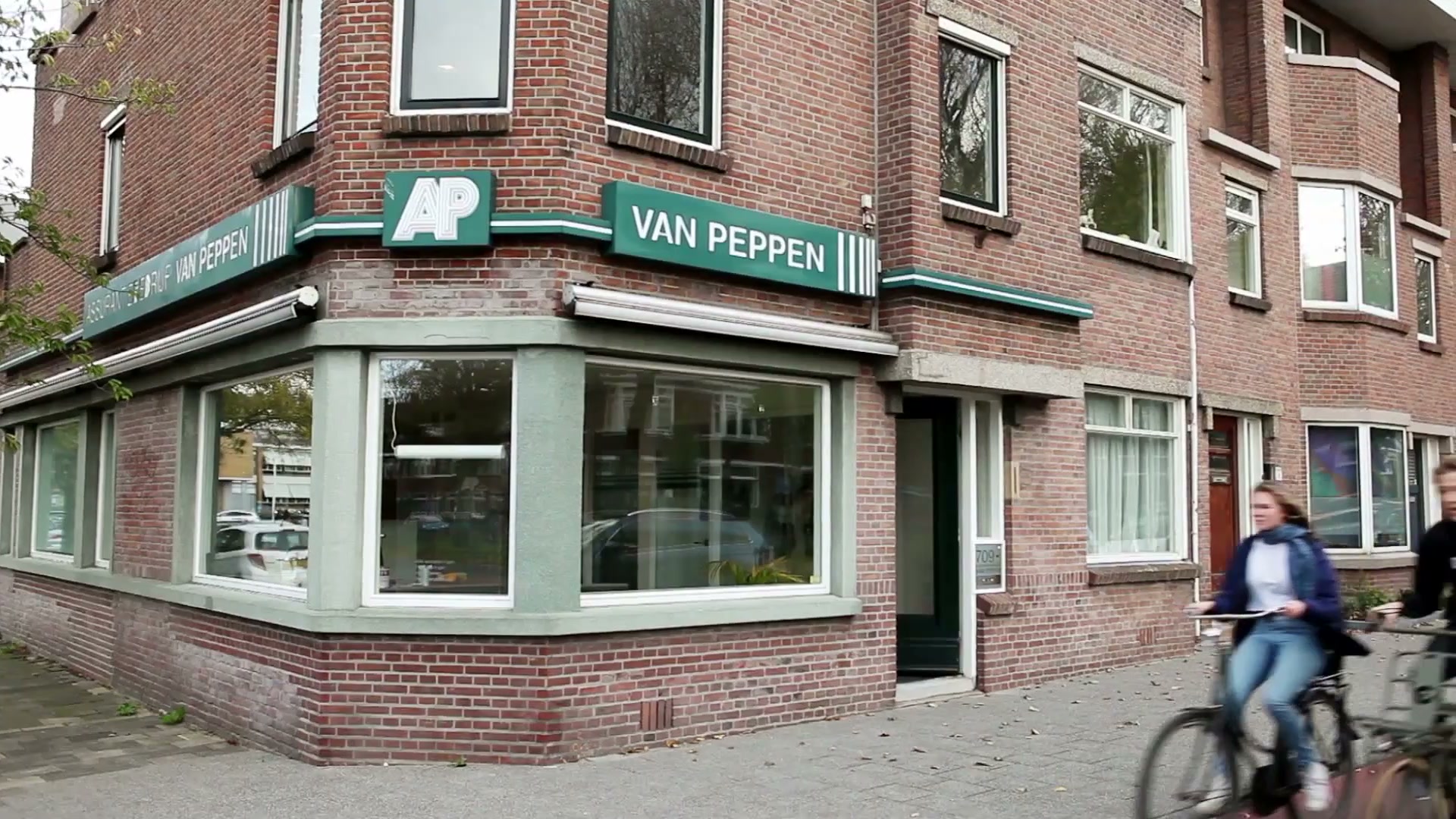 Het hoekpand van Van Peppen met de groene gevelband, het AP-monogram en de letters Van Peppen; op de voorgrond fietst een vrouw voorbij.