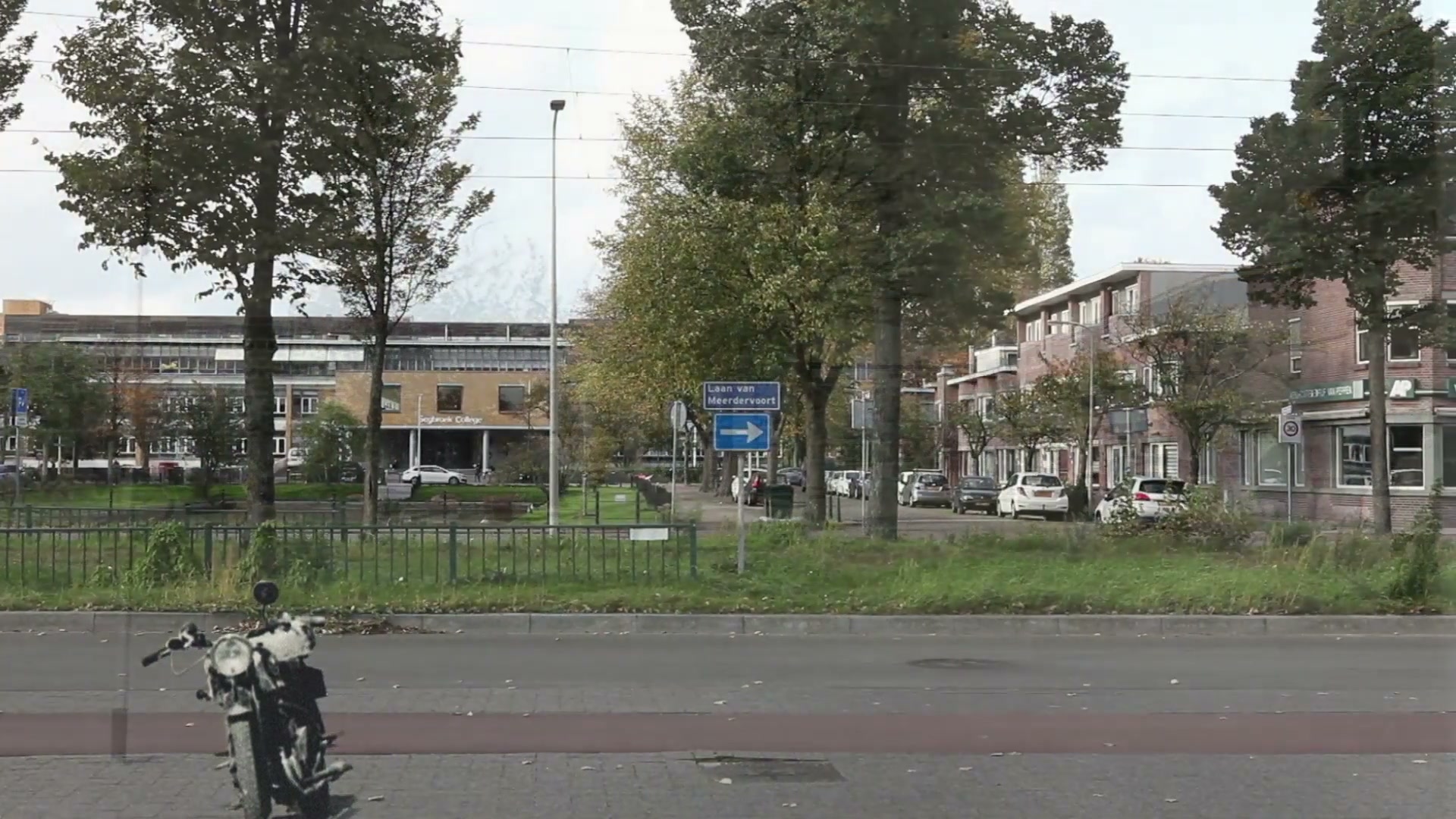 Dezelfde plek in 2017: volgroeide bomen, een hek langs het groen en geparkeerde auto’s. De motor uit de oude foto is in de film als doorzichtig spookbeeld blijven staan.